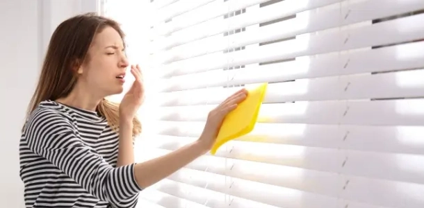 Woman sneezing while dusting window blinds.